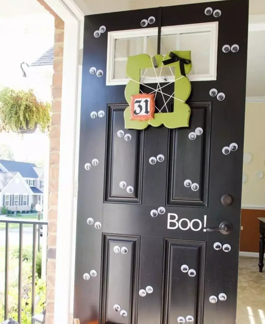 Decoração de porta preta aberta com diversos olhos arregalados, escrita "boo!" e moldura verde escrito "31" com barbante imitando teia e aranhas
