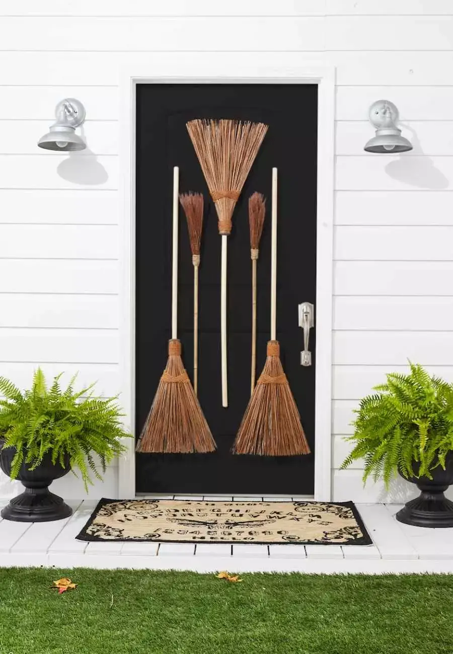 Entrada de casa com parede branca, porta preta com vassouras pregadas e dois vasos de samambaia no chão 