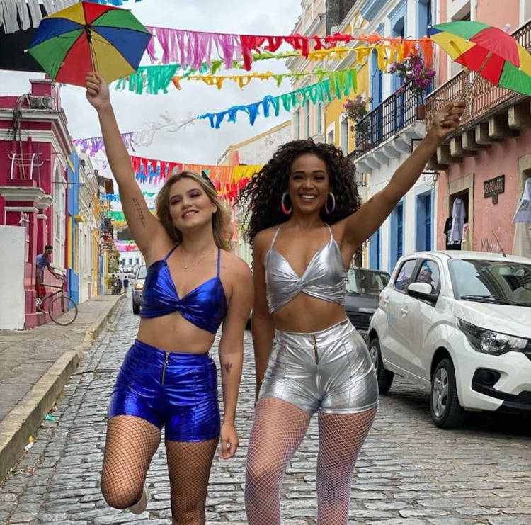 Duas mulheres, uma de pele clara e outra de pele negra, usando conjunto de top e shorts metálicos, sendo um azul e outro prata
