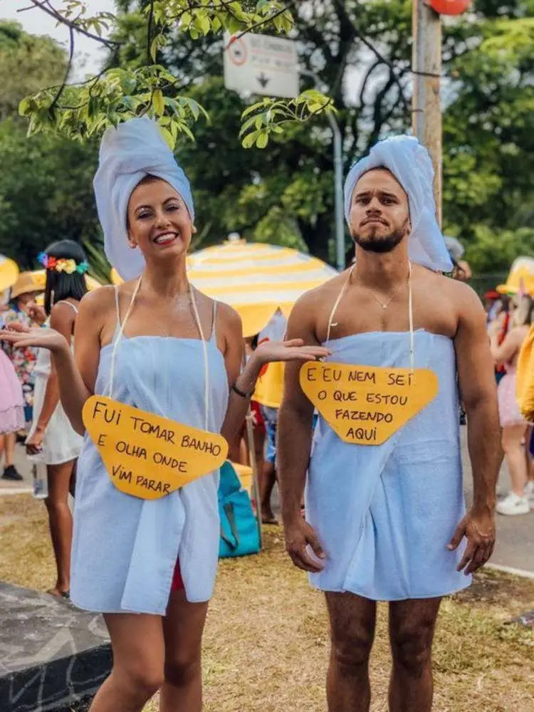 Mulher e homem de pele clara usando toalha enrolada no corpo e na cabeça, com plaquinhas "fui tomar banho e olha onde vim parar" e "e eu nem sei o que estou fazendo aqui"