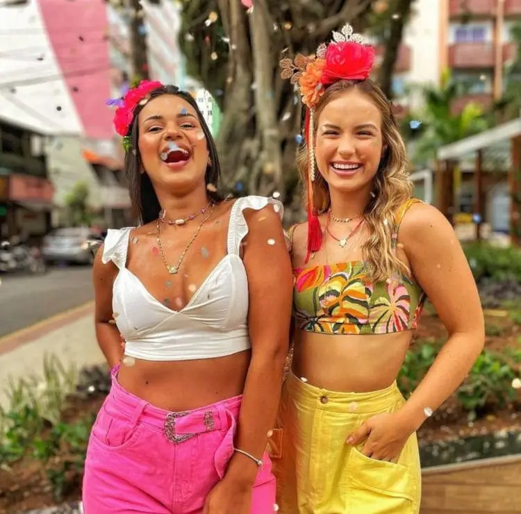 Duas mulheres, sendo uma de pele morena com top branco e shorts rosa e outra de pele clara com top verde de flores e shorts amarelo