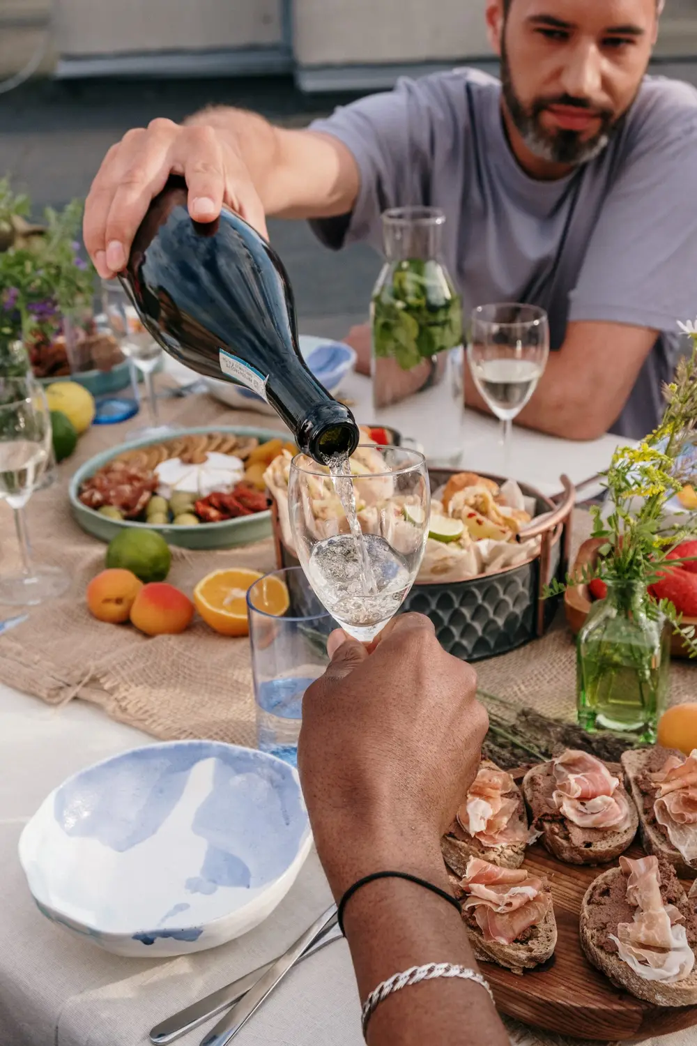 Foto com uma mesa de aperitivos com presento, queijos e duas pessoas bebendo vinho