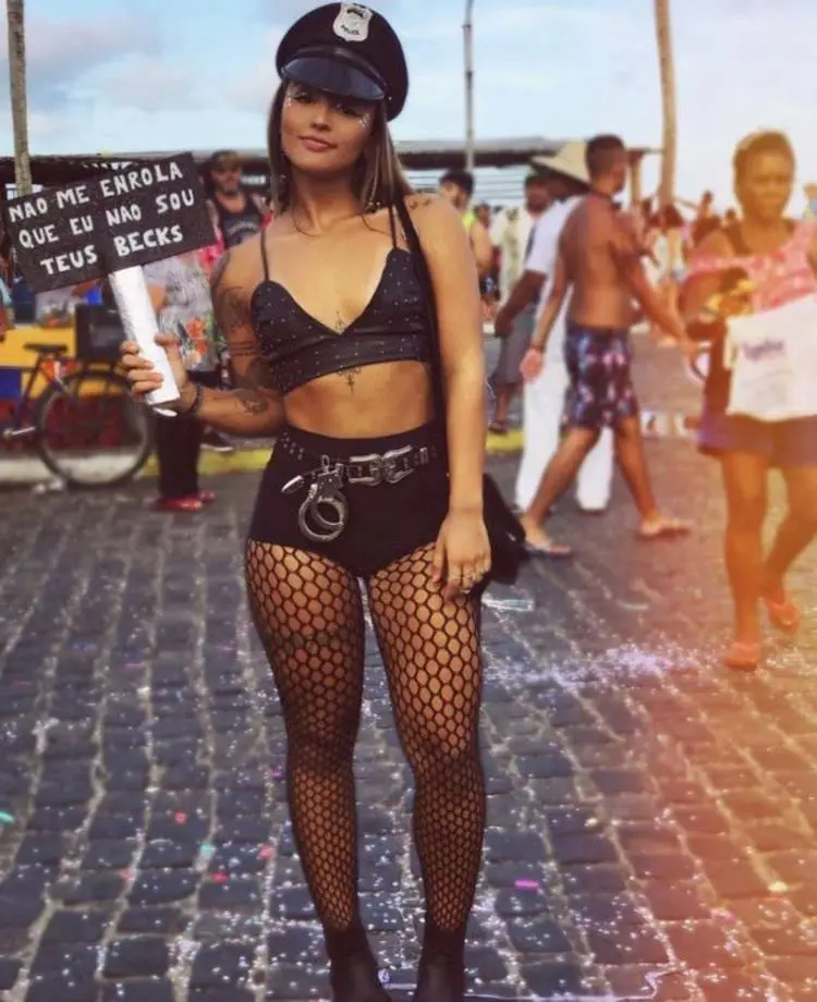 Mulher de pele clara usando look preto composto por meia-calça arrastão, top e hot pant, além de cinto com algema, chapéu de policial e plaquinha escrito "não me enrola que eu não sou teus becks"