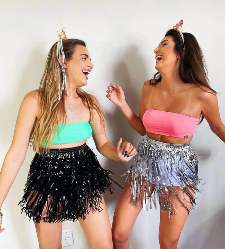 Duas mulheres de pele clara com top e saia com franjas, uma com top verde e saia preta e outra com top rosa e saia prata 