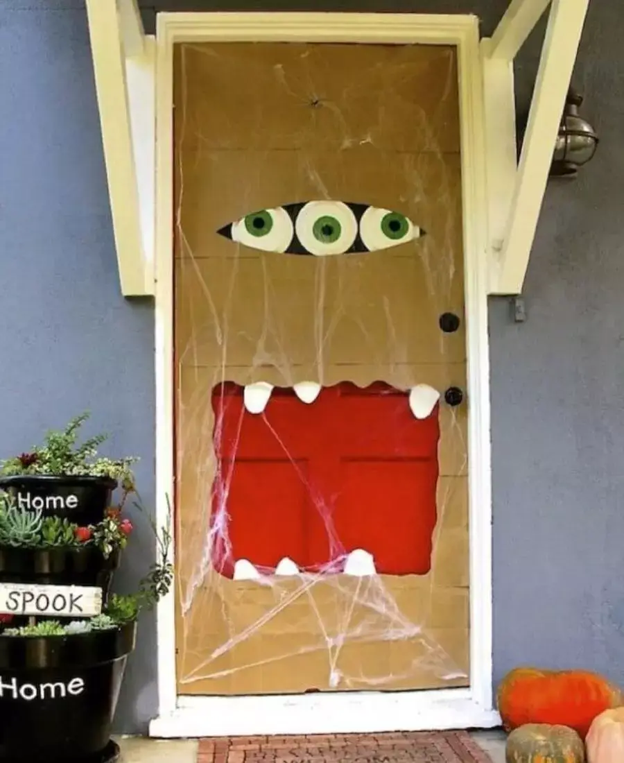 Entrada de casa com parede cinza e porta mrrom com 3 olhos, teias, aranhas e boca de monstro 
