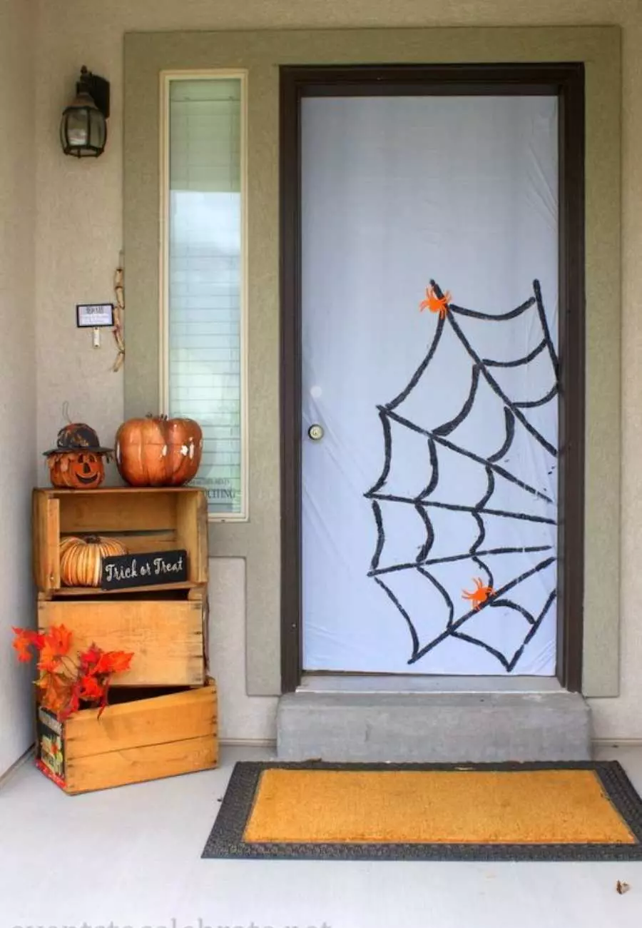 Porta com toalha de plástico branca, teia de aranha desenhada e aranhas de papel. No canto, caixotes de feira com abóboras e flores.