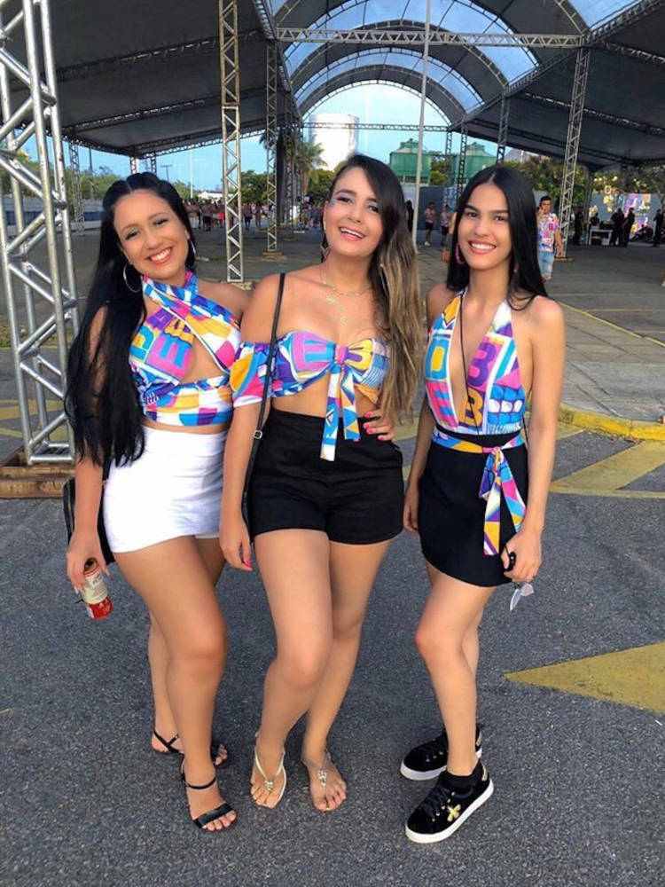 Três mulheres de pele clara com abadá colorido personalizado em tops, sendo que a primeira usa saia branca, a segunda shorts preto e a terceira saia preta