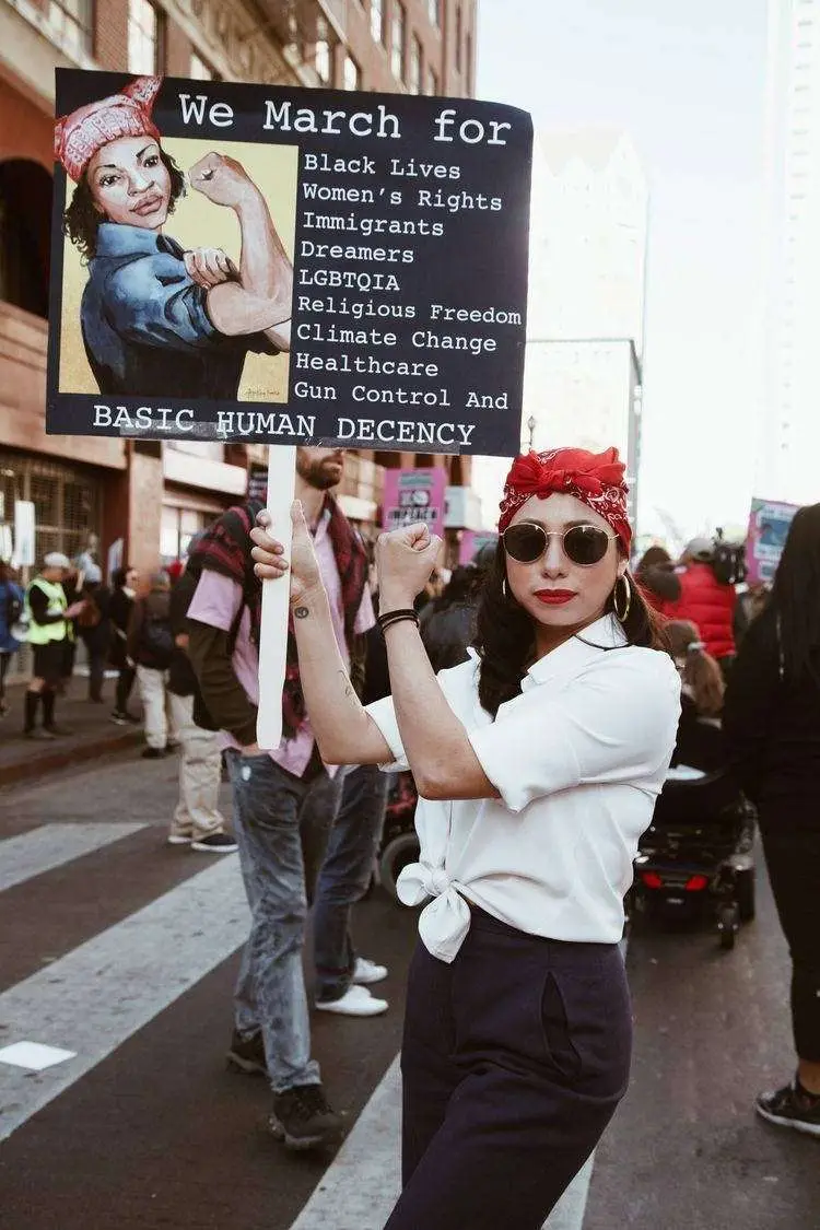 Mulher de pele clara usando lenço vermelho na cabeça, óculos de sol, camisa branca, calça preta, em passeata, segurando cartaz sobre direitos humanos