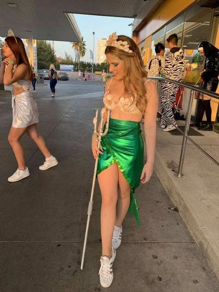 Mulher de pele clara com top bege com pedraria, saia verde imitando cauda, tridente e coroa de conchas