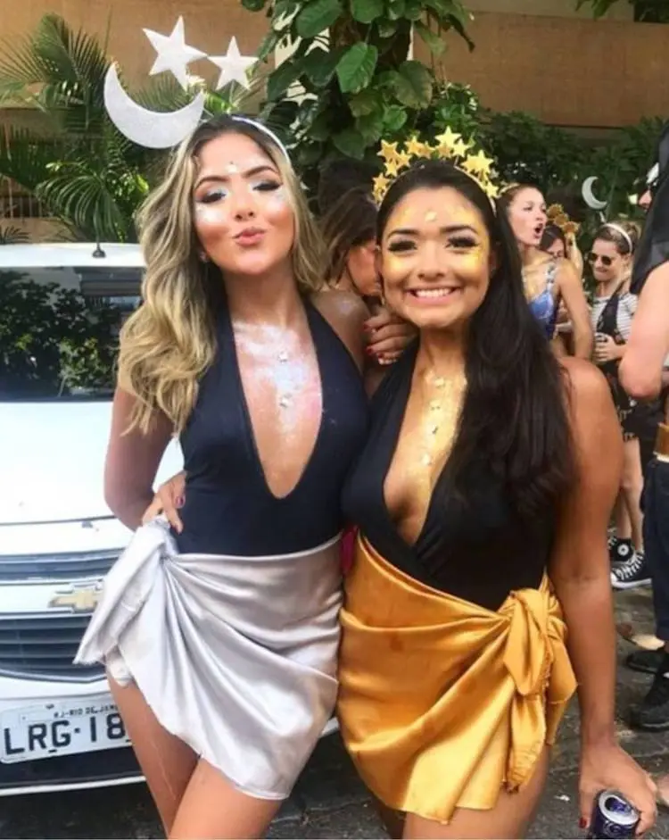 Duas mulheres de pele clara com body preto, sendo uma com saia prata e tiara de lua e outra de saia dourada e tiara de estrelas