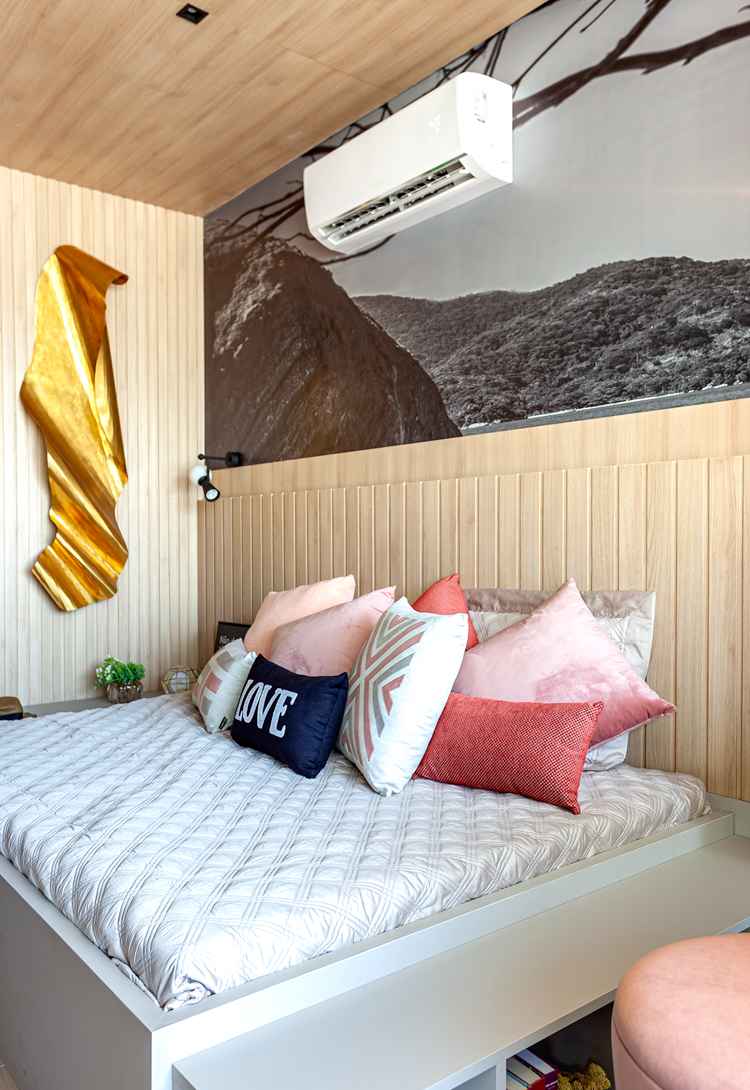 foto de cama de casal com lençol branco, almofadas em rosa, cabeceira de madeira, papel de parede de paisagem e decoração dourada na parede