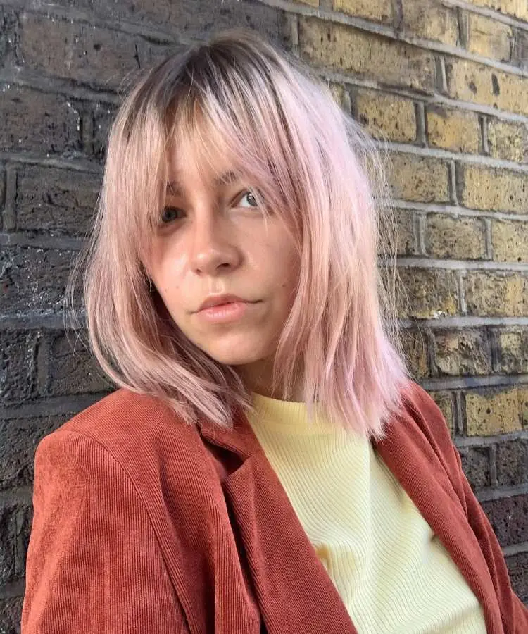 Mulher com cabelo médio em tom pastel rosado, corte reto e franja desfiada. Ela veste blazer laranja e blusa amarela, posando em frente a uma parede de tijolos, com expressão séria e luz natural suave.