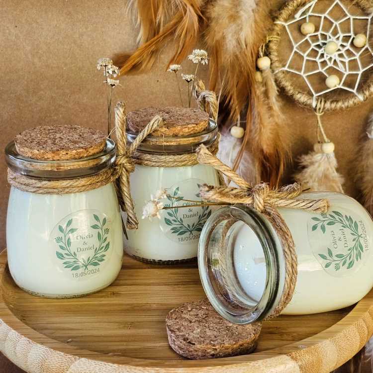 Potes decorativos de vidro com tampas de cortiça, contendo velas brancas. Laços de corda e flores secas adornam os frascos, exibindo etiquetas personalizadas. Fundo rústico inclui filtro dos sonhos com penas e elementos boho-chic, criando atmosfera harmoniosa.