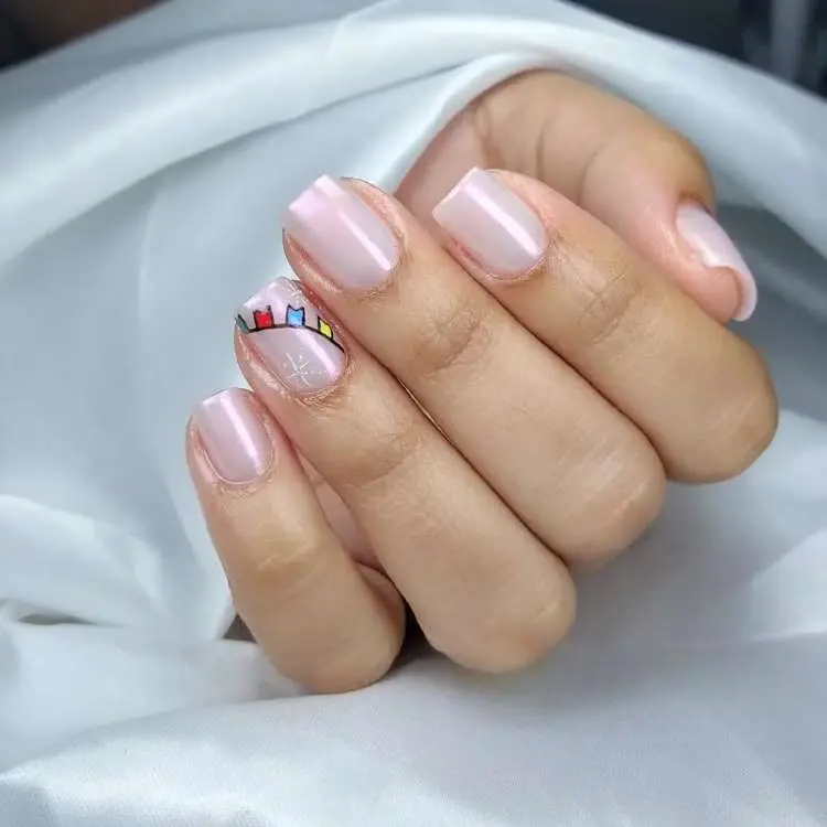 Mão com unhas quadradas e brilhantes em tom nude-rosado. Uma unha tem um desenho de bandeirinhas coloridas e um pequeno brilho em forma de estrela branca. O fundo é um tecido branco.