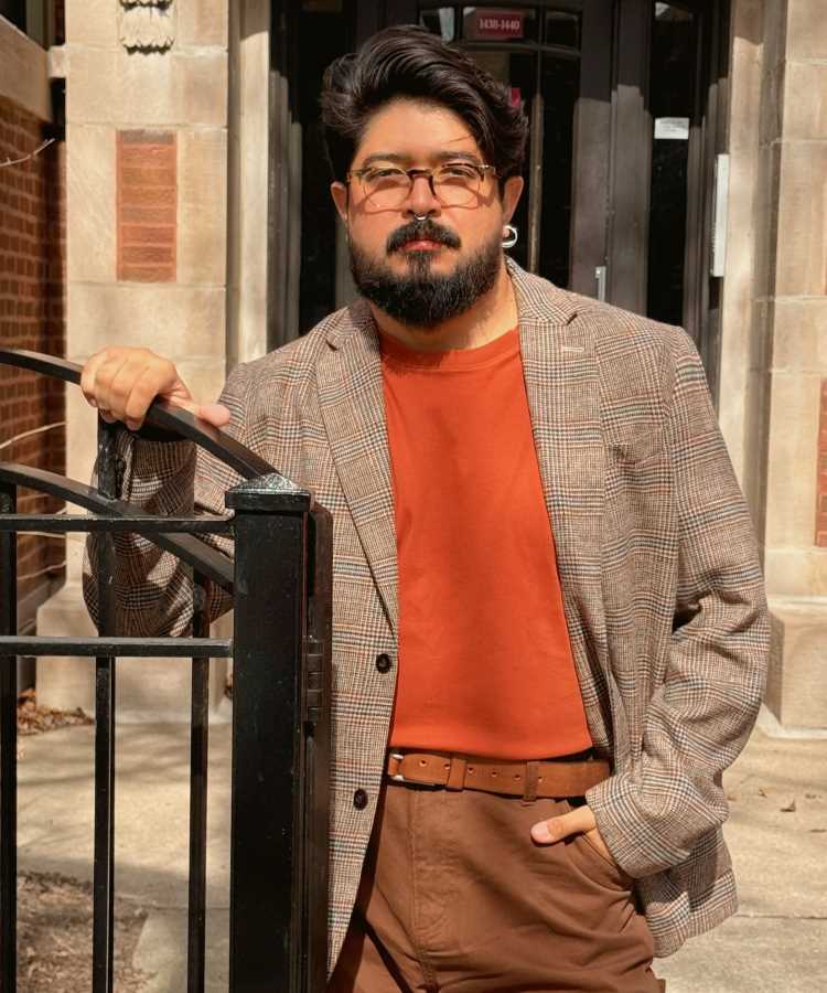Yan Yoshimitsu Sato com barba, cabelos escuros e óculos posa confiante em frente a um portão preto. Ele veste blazer xadrez, camiseta laranja e calça marrom, destacando um estilo elegante urbano em ambiente de tijolos aparentes e portão decorativo.