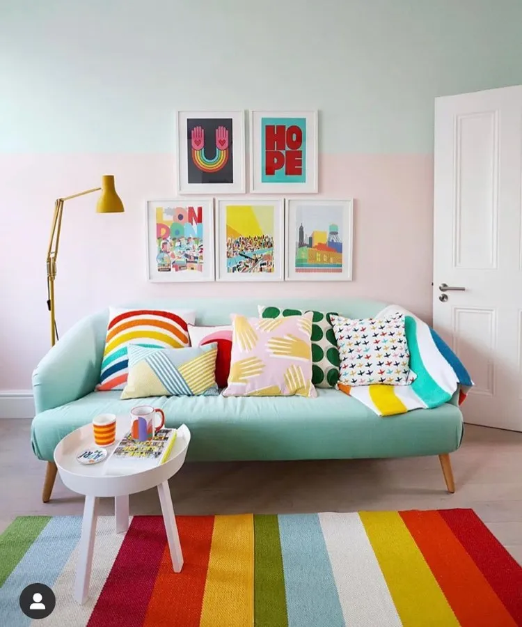 Sala de estar vibrante com sofá azul-claro decorado com almofadas coloridas, mesa lateral branca com canecas, tapete listrado multicolorido e parede adornada por quadros artísticos. Luminária amarela completa o ambiente alegre e acolhedor. 
