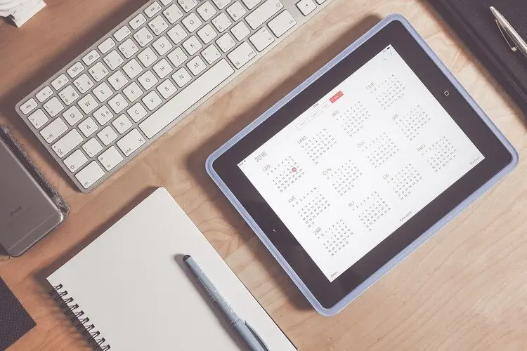 Tablet com um calendário aberto em uma mesa com caderno, caneta e teclado de um computador