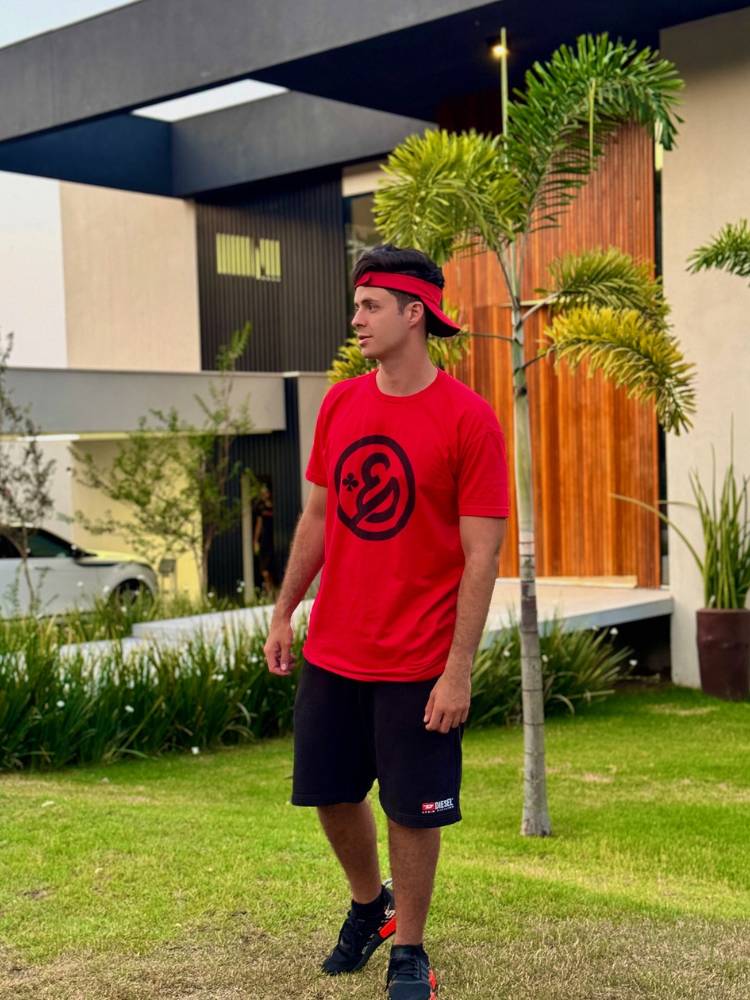 Foto de Enaldinho usando blusa vermelha da sua marca, bermuda preta, em frente a sua mansão