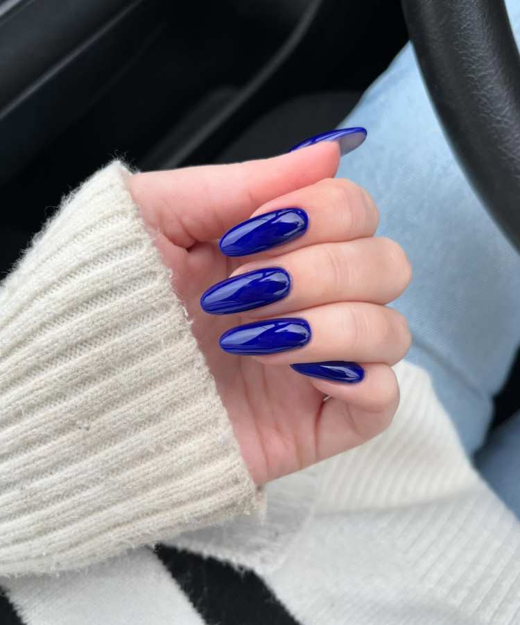 Mão feminina com unhas no formato amendoado, pintadas em tom azul profundo e brilhante. O fundo do carro e a manga de tricô branco criam contraste, destacando a sofisticação dessa cor moderna e elegante, tendência de esmaltes para 2025.