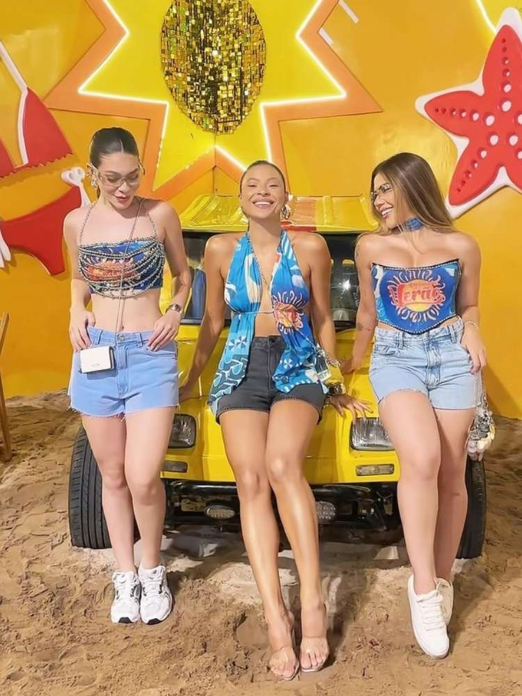 Três mulheres de pele clara em cima de carro amarelo usando abadá azul customizado 