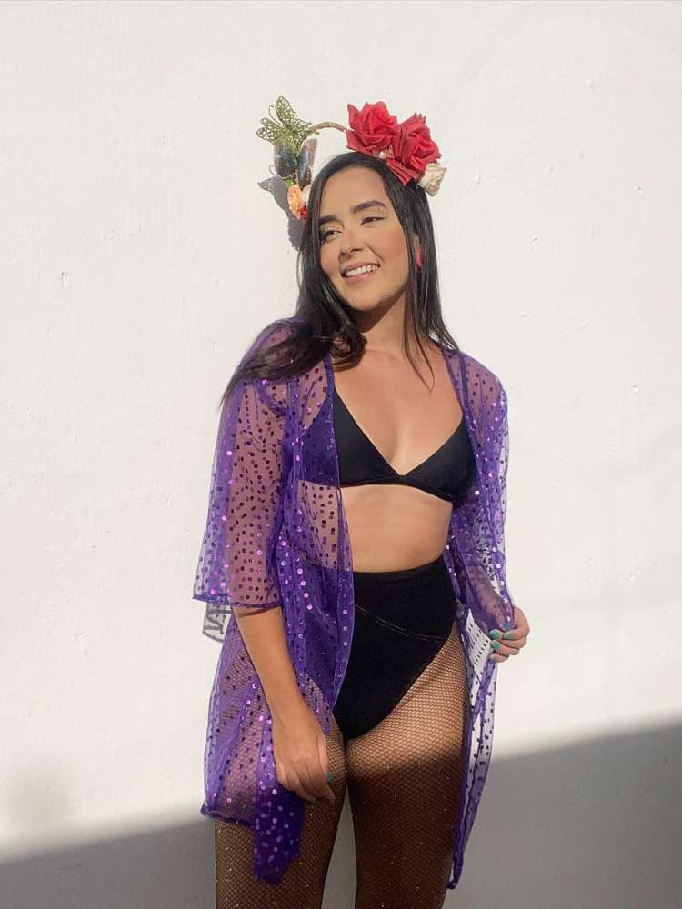 Mulher de pele clara e cabelo preto usando biquíni preto, hot pant preta, sobreposição roxa de brilho e tiara com flores e borboleta. 