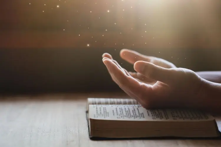 Duas mãos estão estendidas em oração sobre uma Bíblia aberta, cercadas por uma luz suave e partículas brilhantes. A cena transmite espiritualidade, fé e serenidade, com um fundo desfocado destacando a conexão com o divino e a introspecção.