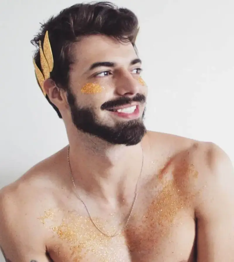 Homem de pele clara com glitter no rosto e ramos em eva dourado no cabelo