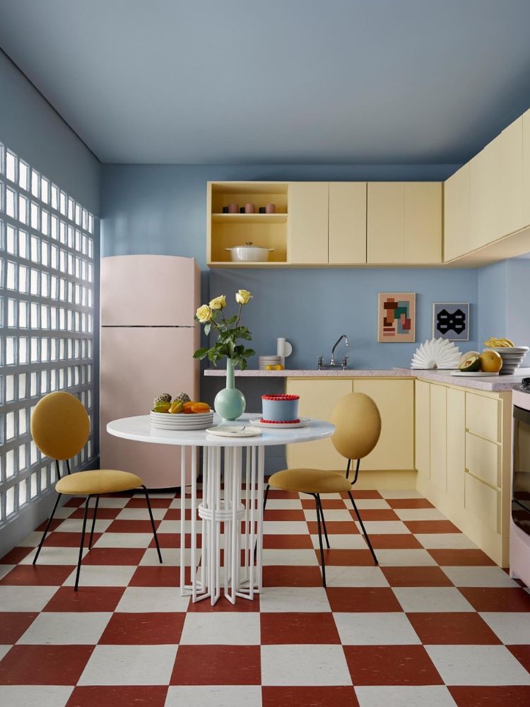 Cozinha colorida retrô em tons pastéis, com parede azul, móveis amarelo-manteiga, chão quadriculado e terracota e geladeira rosa 