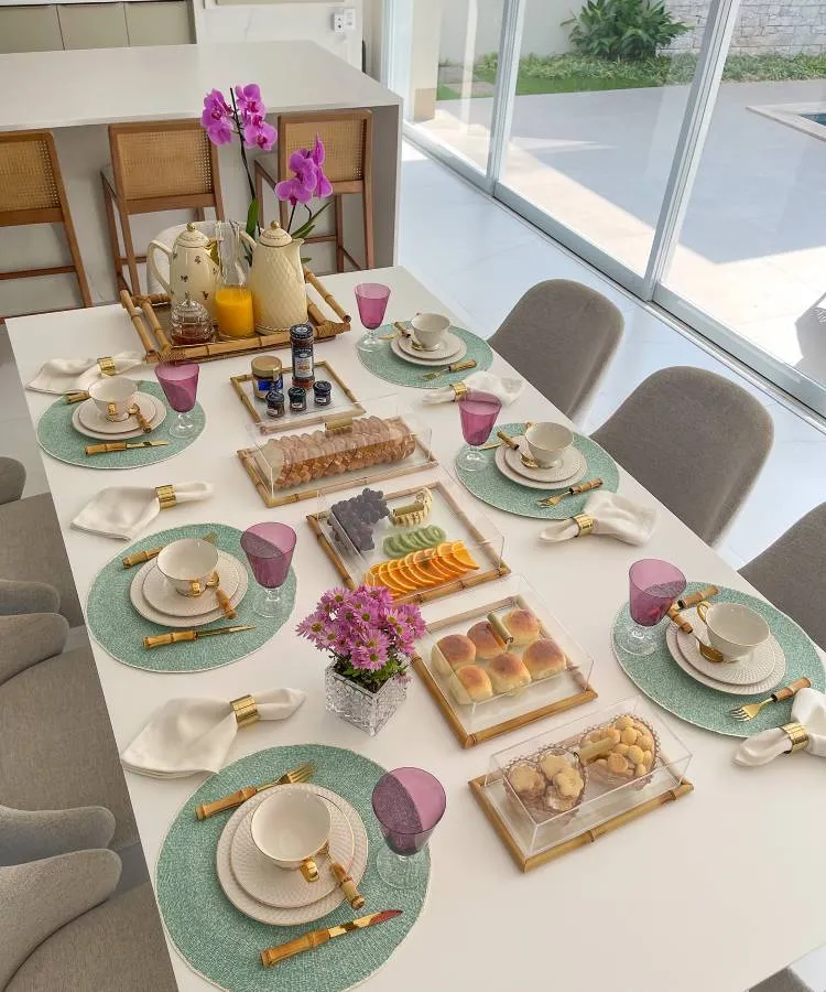A imagem mostra uma mesa posta para café da manhã em um ambiente iluminado. Há sousplats verdes sob pratos brancos e xícaras com detalhes dourados. Talheres de bambu e guardanapos brancos com anéis dourados complementam a arrumação. Copos roxos adicionam um toque de cor. No centro, bandejas contêm pães, biscoitos, frios e frutas fatiadas. Uma jarra de suco e potes de geleia estão dispostos ao lado de uma cafeteira. Um vaso com flores roxas decora a mesa, criando um ambiente acolhedor e elegante.


