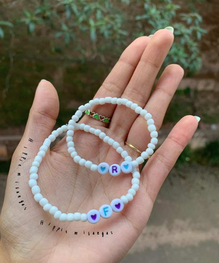 Mão segurando pulseira de miçangas branca, azul e roxo com letra inicial de casal e corações