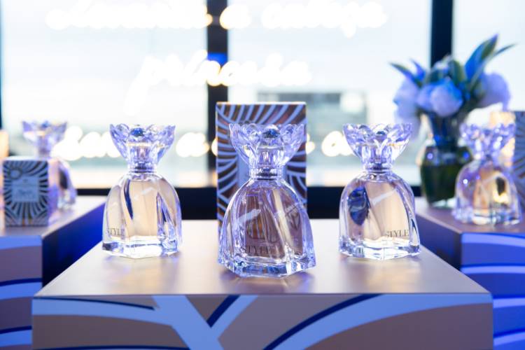 Frascos de perfume cristalinos com tampas decorativas estão expostos em suportes com design azul e dourado. Ao fundo, embalagens combinando e um vaso de flores. A iluminação cria uma atmosfera elegante e sofisticada, destacando o produto no evento.