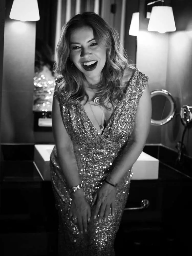 Mulher não-modelo com mais de 40 anos em ensaio fotográfico, usando vestido de paetês dourado, em banheiro. Foto preto e branco