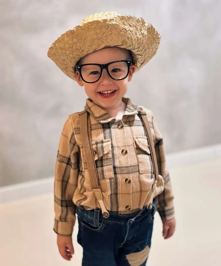 Menino usando chapéu de palha, óculos, blusa xadrez, calça jeans com suspensório.
