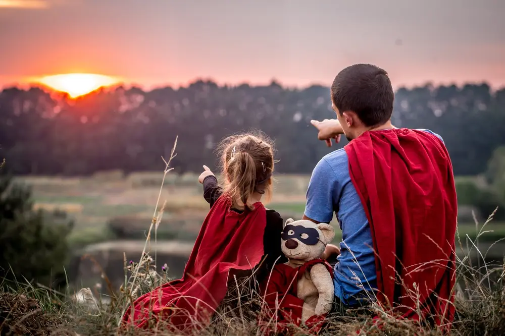 Foto de pai e filha de costas, usando capa vermelha de super-herói e apontando para pôr do sol