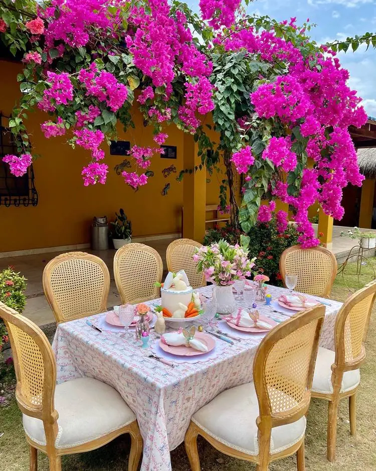  mesa posta de Páscoa para o almoço ao ar livre em casa com planta com flores roxas. A mesa posta contém itens cor-de-rosa e branco
