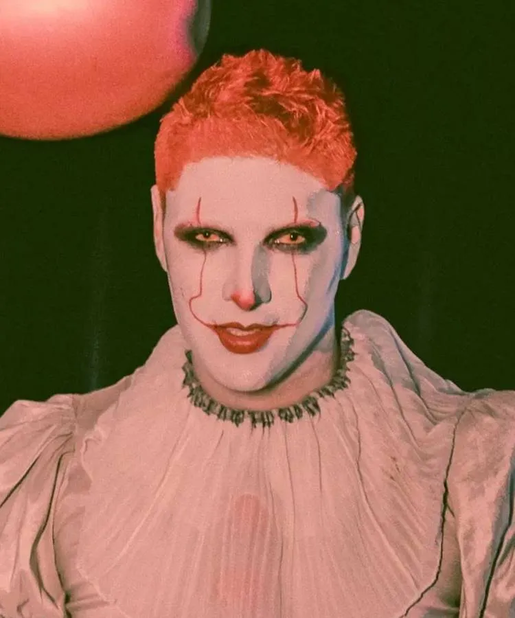 Homem maquiado de Pennywise, com rosto pálido, linhas vermelhas, batom vermelho e fantasia branca 