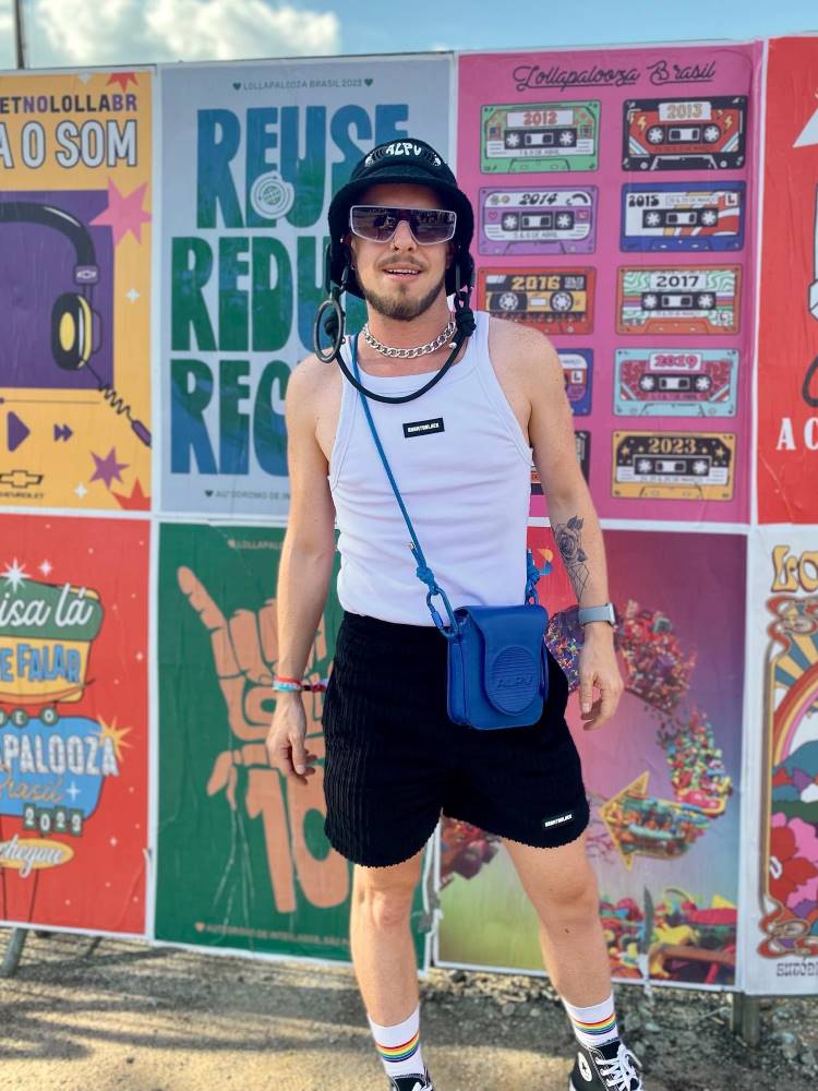 Homem de pele clara no Lollapalooza usando regata branca, bermuda preta, shoulder bag azul, óculos de sol e chapéu preto