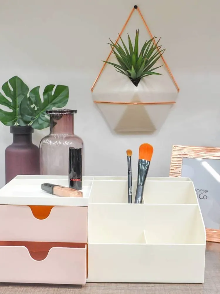 Decoração de uma mesa com um vaso vinho ao fundo com a Monstera, outro vaso transparente, um vaso delicado pendurado e gavetas para organização de maquiagem, além de um porta-retrato.
