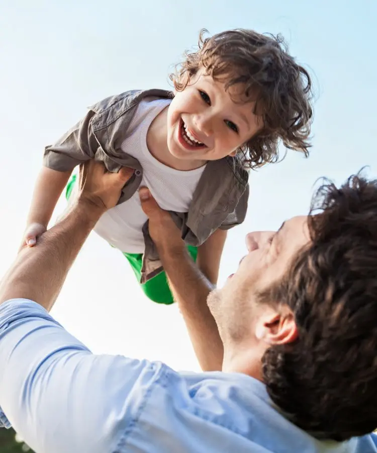 Um pai levanta o filho pequeno no ar, ambos sorrindo com alegria. O menino, com cabelos cacheados, veste camiseta branca, camisa cinza aberta e calça verde. O pai, de baixo, o segura com as mãos estendidas, olhando para cima com um sorriso. O céu azul claro serve de fundo para a cena de felicidade e diversão.