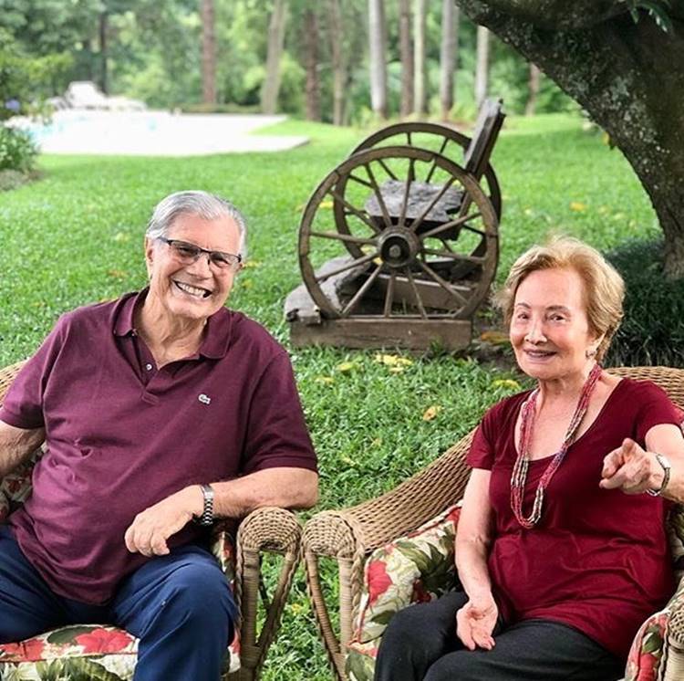 A atriz e o marido que faleceu sentados em cadeira em gramado com árvores