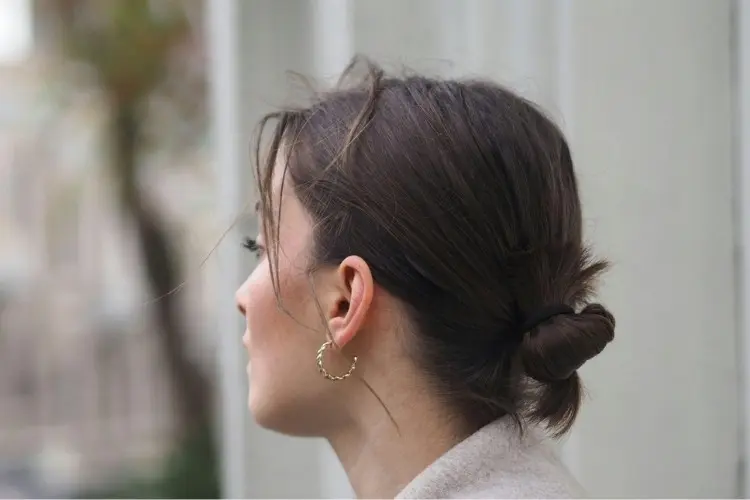 Foto de mulher de perfil. Ela usa o cabelo castanho em um coque baixo e brinco de argola dourado nas orelha