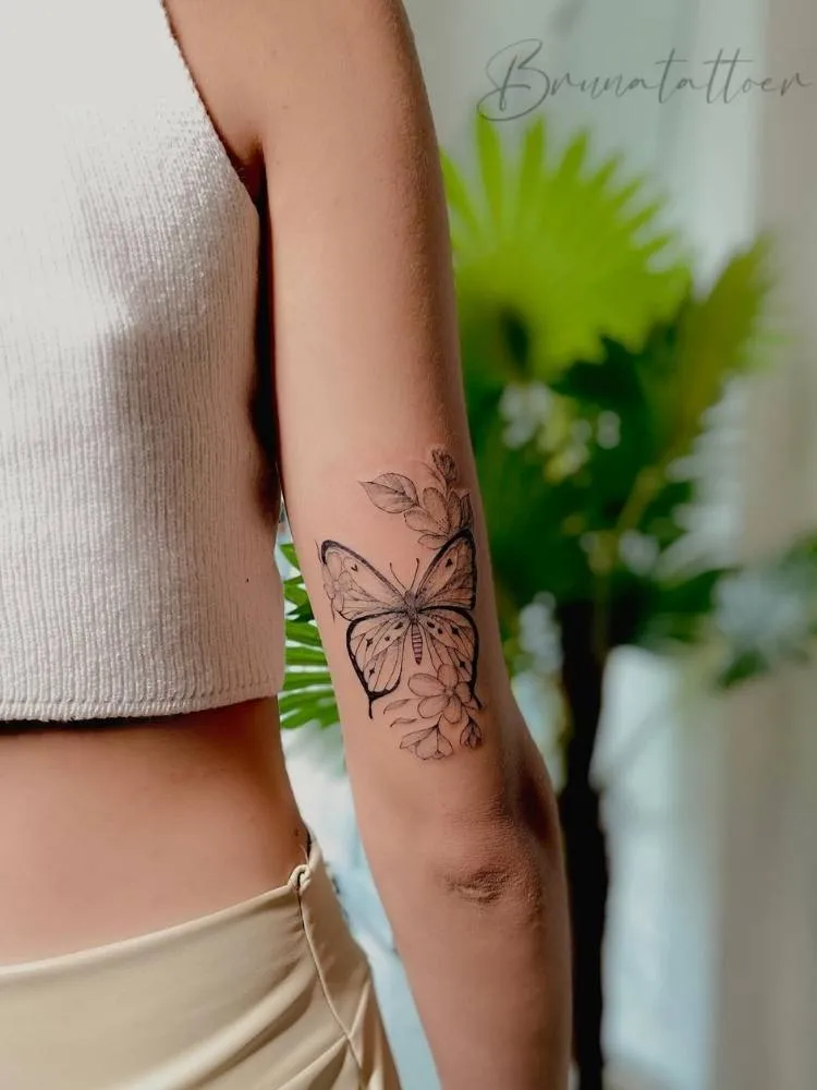 Borboleta delicada com traços finos e mais grossos e flores próximas à ela no braço de uma mulher e pele clara. 
