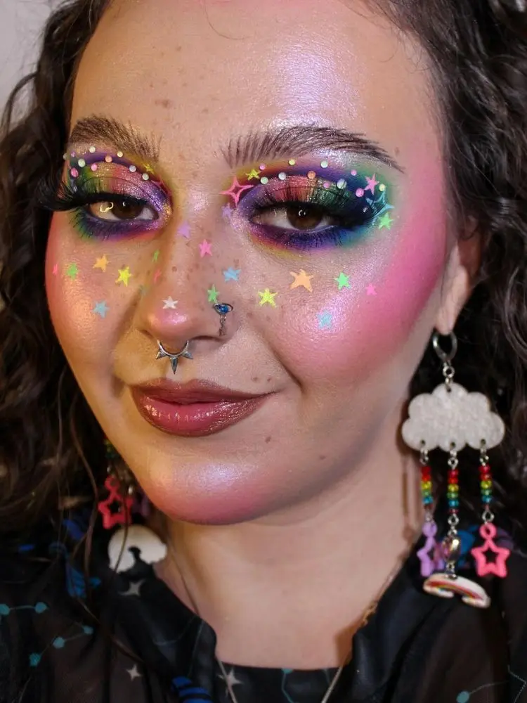 Mulher de pele clara com maquiagem colorida e desenho de estrelas coloridas pelo rosto 