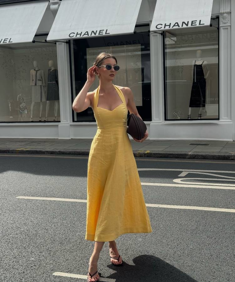 Uma mulher elegante, vestindo um vestido amarelo de alças no pescoço e chinelos de salto, posa na frente de uma loja Chanel. Ela segura uma bolsa grande e escura, ajustando o cabelo com a outra mão.