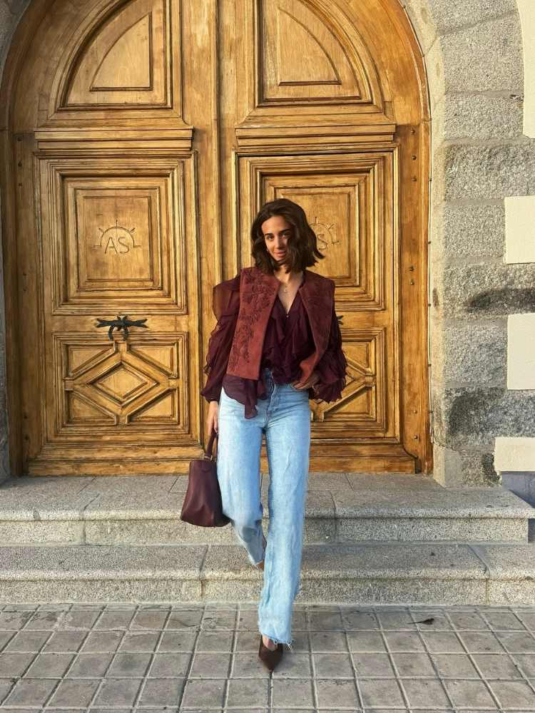 Mulher de pele clara descendo escadas suando camisa marsala com babados, colete bordado, calça jeans e bolsa vinho 