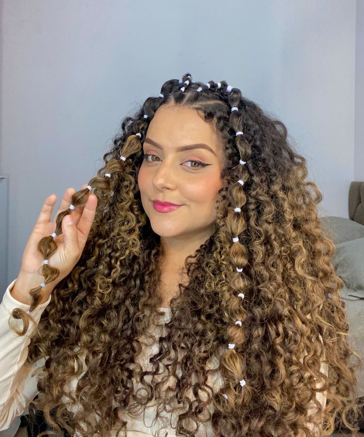 A mulher exibe um penteado estiloso com cachos volumosos e tranças bolha, decoradas com elásticos brancos, que destacam seu cabelo ricamente texturizado. Ela usa maquiagem marcante, com delineador preciso e batom rosa vibrante, sorrindo levemente, transmitindo confiança e estilo moderno.


