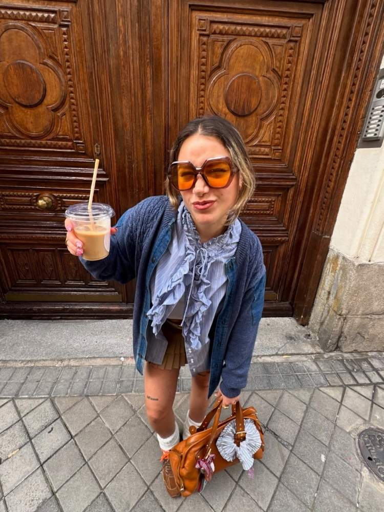 Mulher de pele clara usando óculos de sol oversized, estampado e de lente amarela com camisa azul, jaqueta e saia marrom
