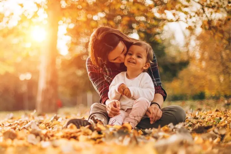 Mãe sentada em um parque outonal cheio de folhas secas, segurando e beijando sua filha pequena que sorri alegremente. A luz suave do pôr do sol ilumina a cena, criando um ambiente acolhedor, cheio de amor e conexão familiar.