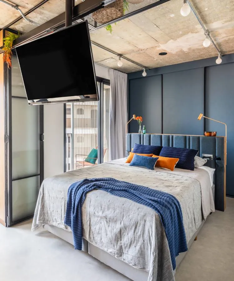 A imagem mostra um quarto moderno. A cama está centralizada, com um edredom cinza e várias almofadas em tons de azul e laranja. Acima da cama, há uma grande TV de tela plana montada no teto, inclinada para baixo em direção à cama para facilitar a visualização. À direita da cama, há uma luminária de chão alta com acabamento dourado, adicionando um toque de elegância ao ambiente. O quarto tem teto de concreto com dutos expostos, conferindo um estilo industrial, enquanto as paredes são pintadas em um tom calmante de azul.
À esquerda da cama, há uma porta de vidro que parece levar a outro cômodo ou talvez a uma varanda, permitindo a entrada de luz natural. De um lado dessa porta de vidro, há plantas em vasos penduradas no teto, introduzindo um toque de verde ao espaço.


