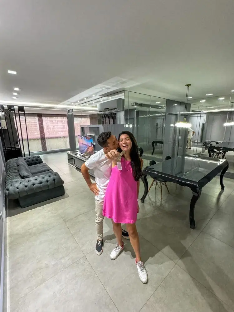 Lara Santana de vestido curto rosa mostrando chave de apartamento, sendo beijada por homem de roupa branca, em apartamento grande 